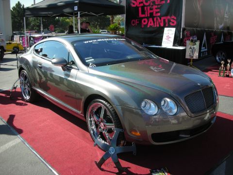 1998 Ford Crown Victoria - gray Bentley Continental coupe sits on a red carpet display beneath a Secret Life of Paints banner at an indoor show.