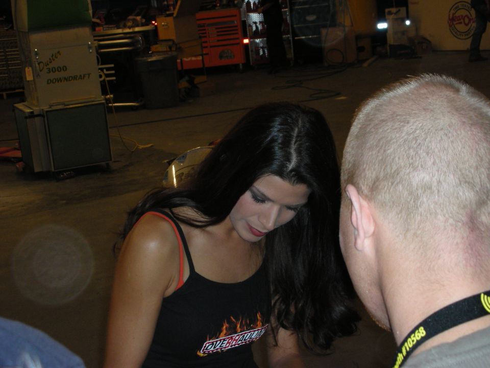 Dark-haired woman in flame-logo tank top beside a shaved-head man in a shop