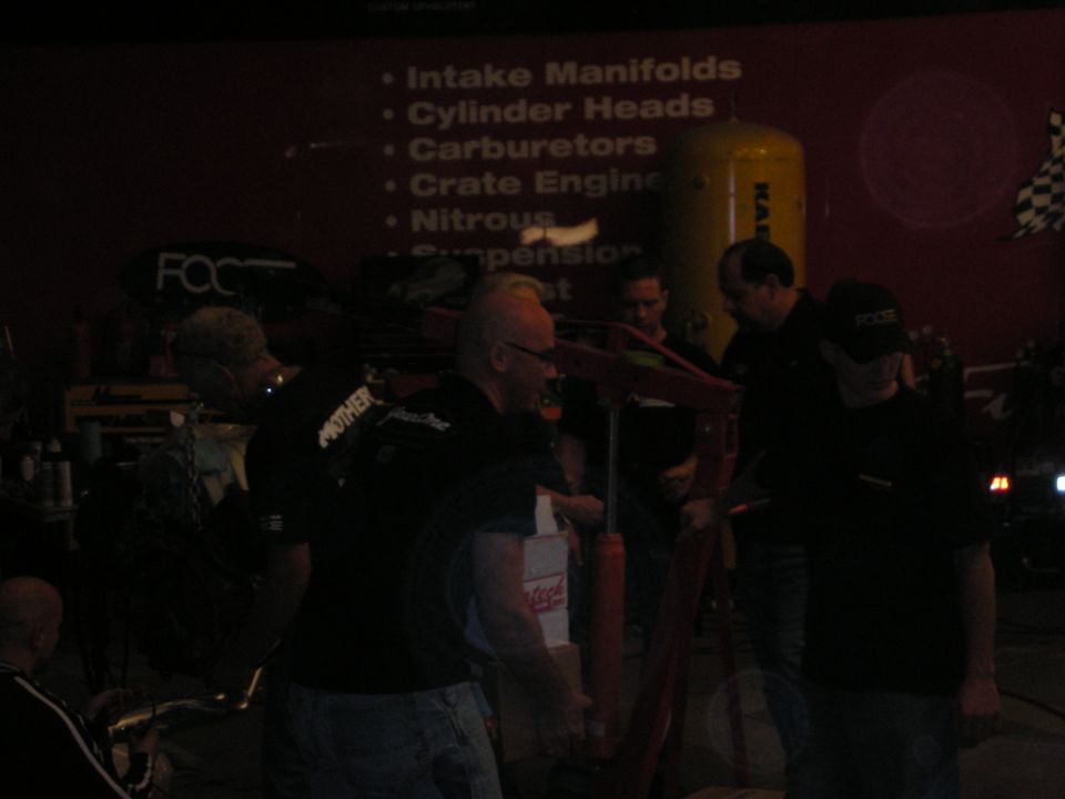 Dim shop scene with men gathered before a red banner listing engine parts