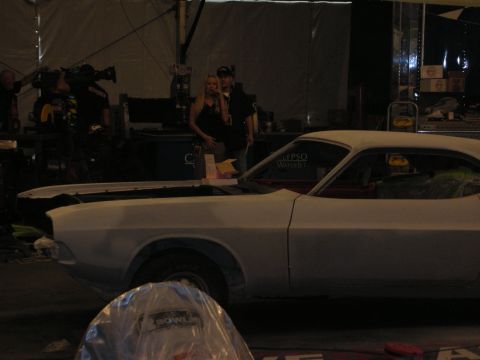 1998 Ford Crown Victoria - white primered muscle car sits in a dim work tent as a blond woman and crew with a camera stand behind it.