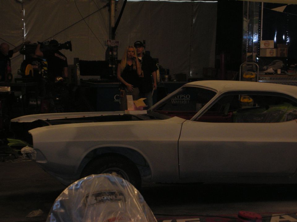 White primered muscle car in a dim tent with crew and camera in background