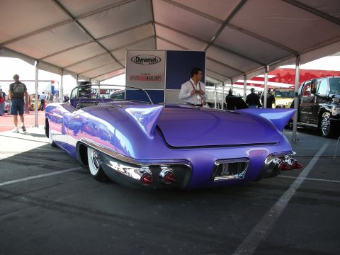 1998 Ford Crown Victoria - finned rear of a purple custom convertible faces a Dynamat and Overhaulin' sign as a man stands beside it under a tent.
