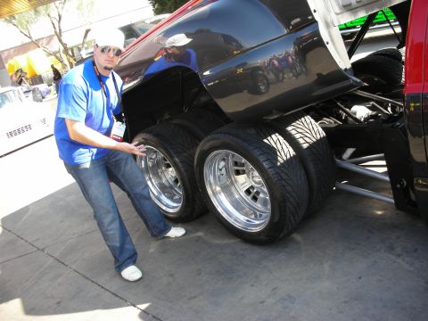 1998 Ford Crown Victoria - man in a blue polo and white cap touches the dual chrome-wheeled rear tires of a lifted black truck at an outdoor show.