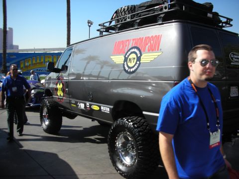 1998 Ford Crown Victoria - lifted gray cargo van with Mickey Thompson tire graphics and a roof rack on display, a man in a blue shirt walking past.