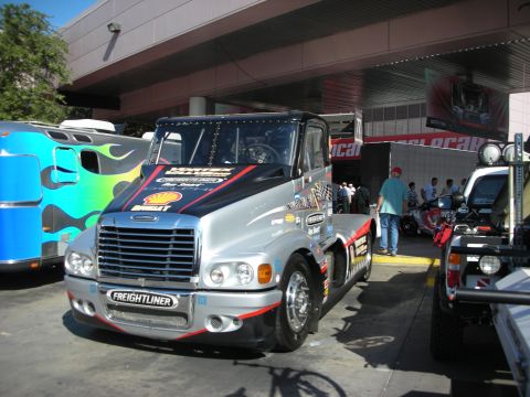 1998 Ford Crown Victoria - customized silver and black Freightliner semi-truck cab with sponsor decals parked under an awning beside a flame-painted bus.