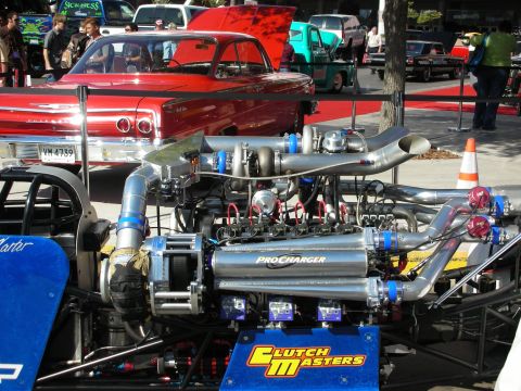 1998 Ford Crown Victoria - chrome ProCharger-supercharged dragster engine with red plug wires, displayed near a red classic car at an outdoor event.