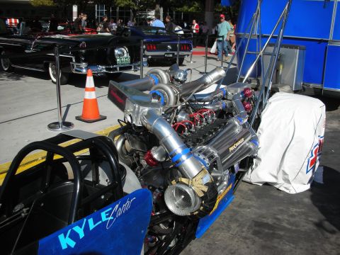 1998 Ford Crown Victoria - chrome twin-turbo ProCharger dragster engine on a blue rail car lettered Kyle Carter, displayed by an orange traffic cone.