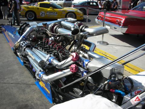 1998 Ford Crown Victoria - polished supercharged dragster engine on a blue chassis, with a yellow number-21 race car and red classic car in the background.
