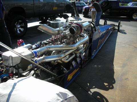 1998 Ford Crown Victoria - chrome turbocharged engine mounted on a long blue dragster rail car parked beside a lifted truck at an outdoor show.