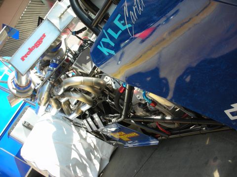 1998 Ford Crown Victoria - Low-angle view of a blue Kyle Carter dragster's chrome engine and stainless headers, with a Prologger banner in the pit area.