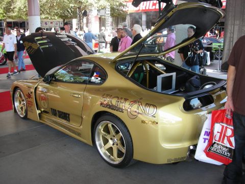 1998 Ford Crown Victoria - gold Toyota Supra with raised hood and hatch, roll cage, and High End and Top Secret graphics, displayed in a crowd.