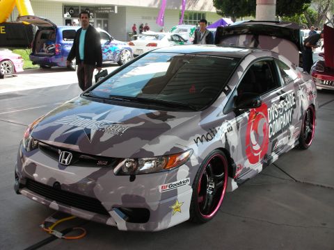 1998 Ford Crown Victoria - Honda Civic Si coupe wrapped in gray urban camouflage with sponsor decals and pink-lipped black wheels at a show.