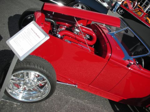 1998 Ford Crown Victoria - bright red open-top hot rod roadster sits at a show with polished exhaust piping, a red engine bay and chrome five-spoke wheels.