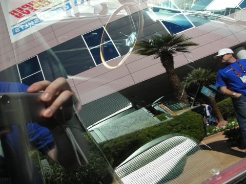 1998 Ford Crown Victoria - Buildings, palm trees and a man in a blue shirt are mirrored in the curved glass of a car at an outdoor event.