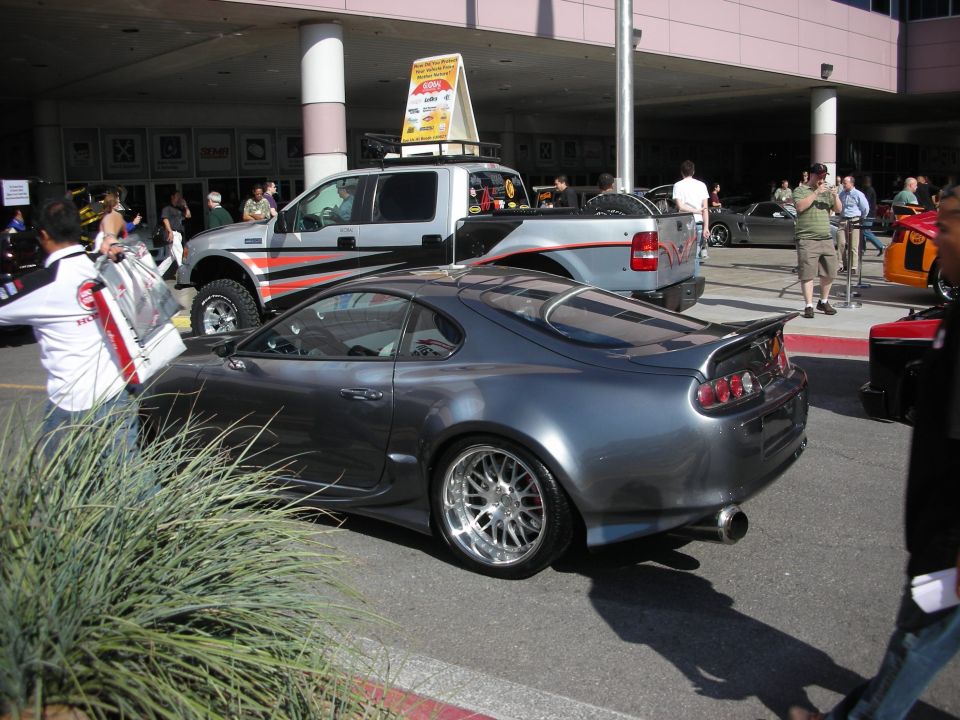 Grey sports coupe with large rear wing parked near a lifted pickup at an outdoor event