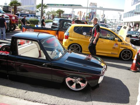 1998 Ford Crown Victoria - slammed dark mini pickup with chrome wheels sits beside a yellow K20-powered hatchback at a sunny outdoor show.