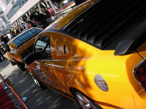 1998 Ford Crown Victoria - glossy yellow modern Mustang's side and rear window louvers shine in the sun, with another yellow Mustang ahead.