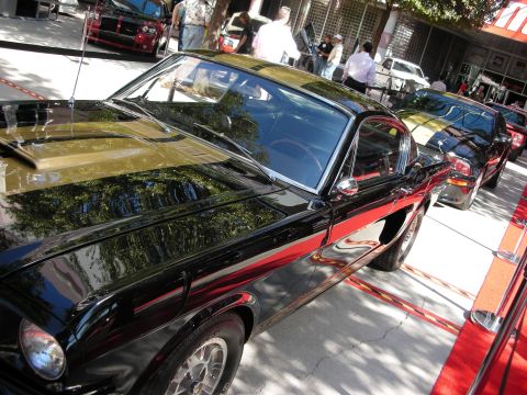 1998 Ford Crown Victoria - glossy black fastback muscle car with a gold racing stripe sits roped off on red carpet at an outdoor show.