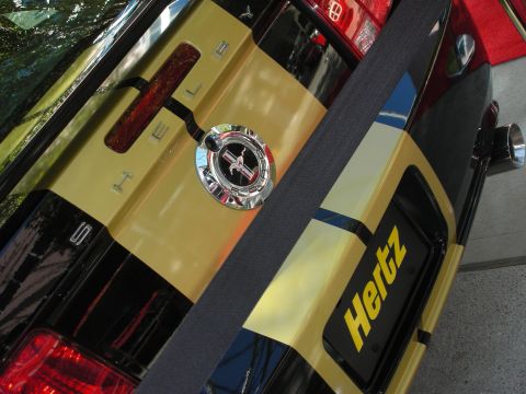 1998 Ford Crown Victoria - Close-up of a yellow and black car's rear deck showing a chrome fuel cap, racing stripe and 'Hertz' lettering.