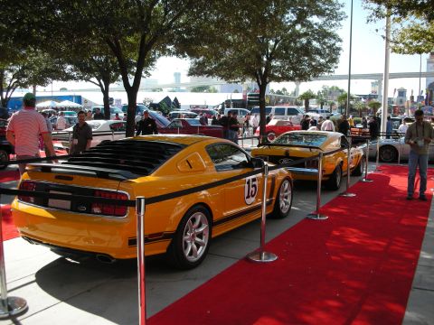 1998 Ford Crown Victoria - yellow coupe marked with number 15 racing livery sits with an orange car on red carpet at a crowded outdoor show.