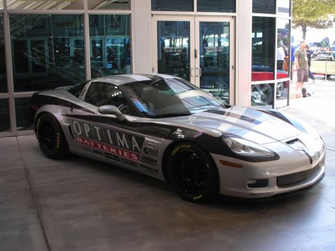 1998 Ford Crown Victoria - silver and black sports coupe wrapped in Optima Batteries sponsor graphics sits on pavement outside a glass-front building.