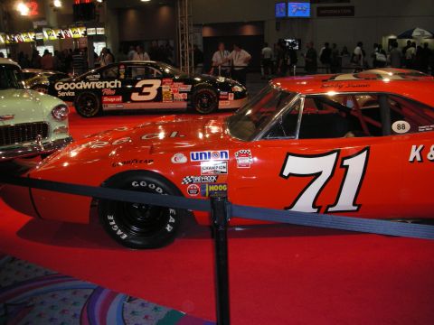 1998 Ford Crown Victoria - orange number 71 vintage stock car displayed beside a black number 3 Goodwrench race car on red carpet indoors.