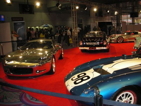 1998 Ford Crown Victoria - mirror-finish chrome concept coupe, a black race car and a blue number 98 racer share an indoor red-carpet display.