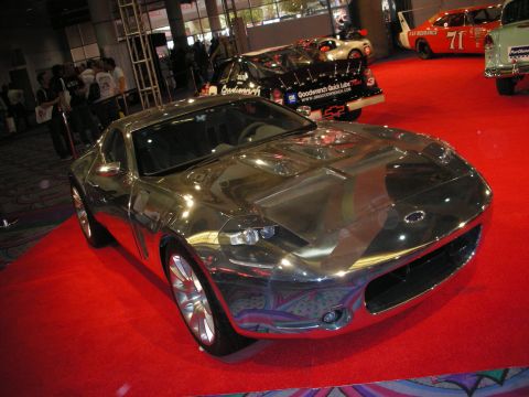 1998 Ford Crown Victoria - mirror-polished chrome concept sports coupe sits front and center on red carpet with race cars in the background.