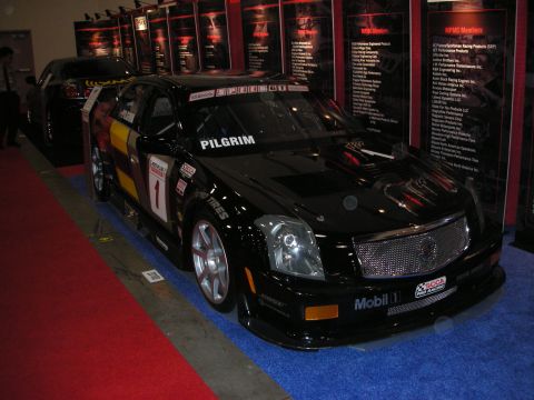 1998 Ford Crown Victoria - black Cadillac CTS race car wearing number 1 and 'Pilgrim' sponsor livery sits on blue carpet at an indoor show.