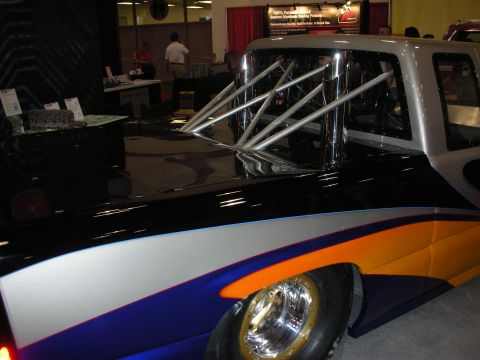 1998 Ford Crown Victoria - Side view of a low black-and-orange race truck body with tubular wheelie-bar framework and wide rear slick on a show floor.