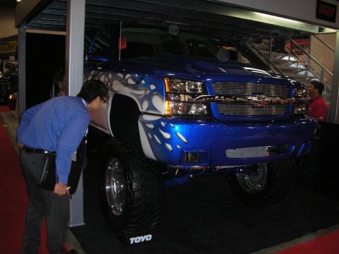1998 Ford Crown Victoria - lifted blue Chevrolet pickup with white flame graphics and large off-road tires on a display stand as a man inspects it.