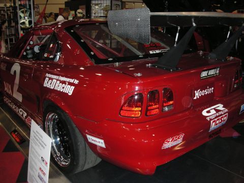 1998 Ford Crown Victoria - Rear-quarter view of a dark red Mustang road-race car with a tall rear wing, number 2 and B&D Racing and Hoosier decals.