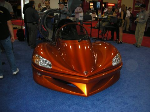 1998 Ford Crown Victoria - Copper-orange teardrop-bodied concept car with a clear bubble canopy displayed on blue carpet among trade-show attendees.