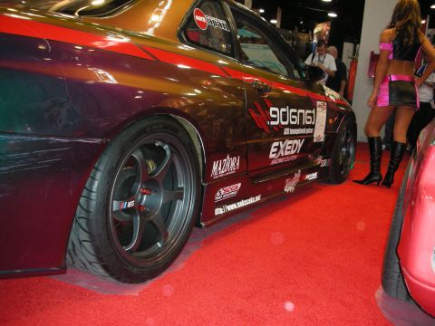 1998 Ford Crown Victoria - Side profile of a multicolor show car with deep-dish wheels and racing decals, beside a model in pink on a red carpet.