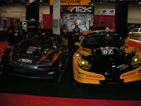 1998 Ford Crown Victoria - black and a yellow sports car with flame and racing graphics displayed side by side at an ARK Performance booth.