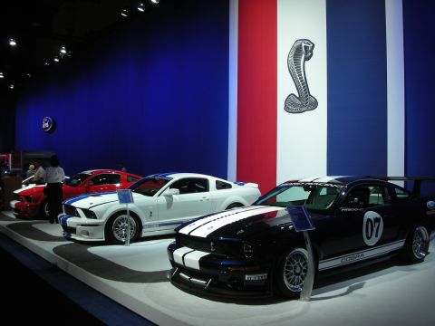 1998 Ford Crown Victoria - row of three Shelby Mustangs in red, white and black-with-stripes beneath a large Cobra snake banner at a show.