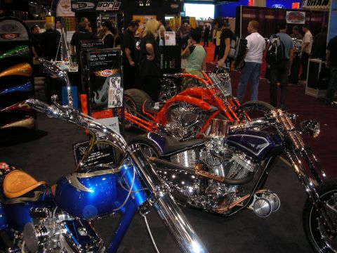 1998 Ford Crown Victoria - row of custom chopper motorcycles with heavy chrome and orange and blue paint at a crowded trade show booth.