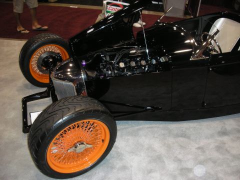 1998 Ford Crown Victoria - black hot rod roadster with chrome grille and orange-painted wire-spoke wheels sits on a polished show-floor display.