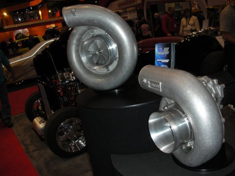 1998 Ford Crown Victoria - Two oversized polished aluminum centrifugal superchargers sit on a black pedestal in front of a hot rod at a trade show.