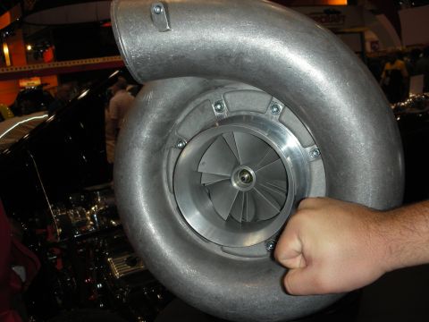 1998 Ford Crown Victoria - hand grips the volute of an enormous polished centrifugal supercharger, its impeller visible, at a trade-show booth.