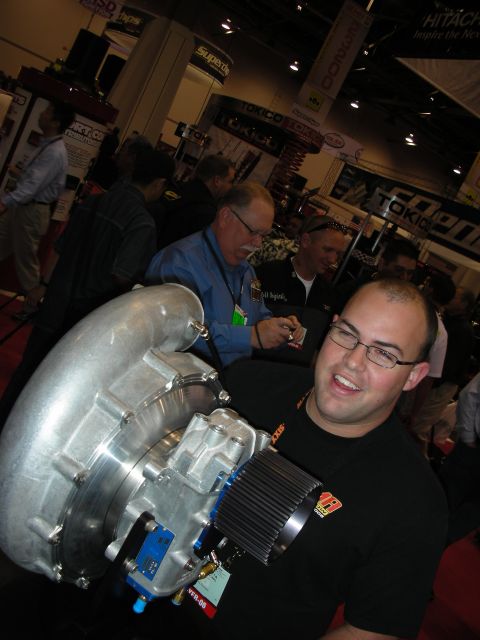 1998 Ford Crown Victoria - grinning bald man in a black shirt holds a large cast-aluminum supercharger unit amid a crowded trade-show aisle.