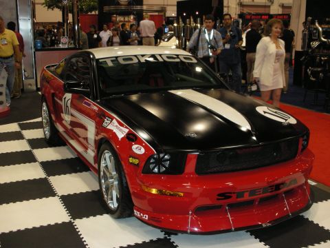 1998 Ford Crown Victoria - red-and-black Steeda-bodied Ford Mustang with race number 10 and Tokico graphics sits on a checkered show-floor.