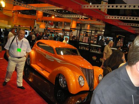 1998 Ford Crown Victoria - orange custom coupe with subtle flame graphics and a chopped roof draws onlookers on a red-carpeted show floor.