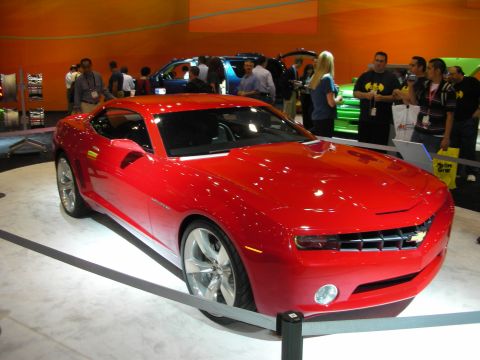 1998 Ford Crown Victoria - red Chevrolet Camaro coupe sits behind stanchions on a white show floor with a crowd milling around it.
