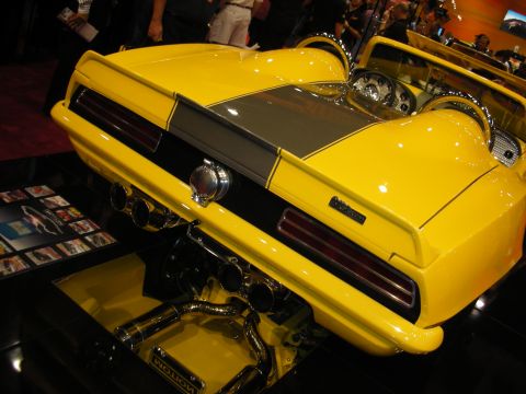 1998 Ford Crown Victoria - rear of a yellow Motion custom car reveals dual exhaust headers and a chrome engine through a cutaway floor display.