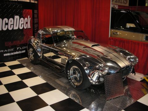 1998 Ford Crown Victoria - bare polished-aluminum Cobra-style roadster sits on a black-and-white checkered floor at a RaceDeck show booth.