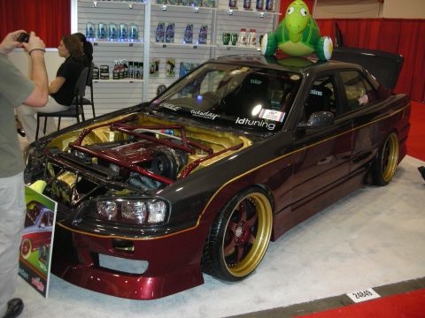 1998 Ford Crown Victoria - slammed maroon JDM sedan with gold wheels, exposed engine bay, and a cartoon turtle mascot on its roof sits on display.