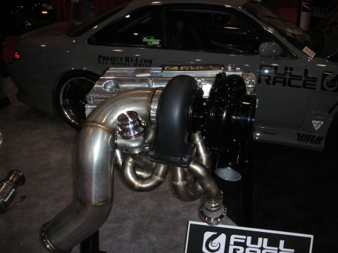 1998 Ford Crown Victoria - large polished turbocharger and exhaust manifold displayed on a stand at a Full Race booth, with a silver coupe behind.