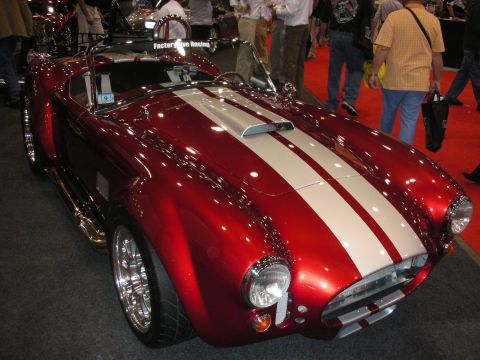 1998 Ford Crown Victoria - candy-red Cobra-style roadster with white racing stripes and chrome wheels, displayed at a Factory Five Racing booth.
