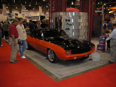 1998 Ford Crown Victoria - black-and-orange restomod first-generation Camaro on a red carpet, drawing spectators at an indoor trade show.